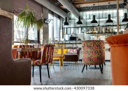 Charming hall in a loft style in a mexican restaurant with open kitchen on the background. In front of the kitchen there are wooden tables with multi-colored chairs and sofas. On the sofas there are