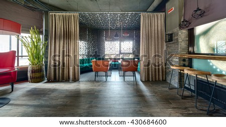 Room in a loft style in a mexican restaurant. On the left there is a crimson armchair with a glass table near it and a red lampshade over it. Big cactus in a wicker pot is behind the armchair. On the