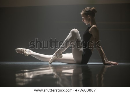 Pretty ballerina sitting on the floor in the dance hall. She leans her hands on the floor from the back while pulling one leg forward and the other bent at the knee. She is reflected on the floor