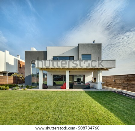 Beautiful country house in a modern style with the large lawn. In front of the house there is a covered terrace with a lounge zone and a furnace. On the lawn there is a tree and flowerbeds. Outside.