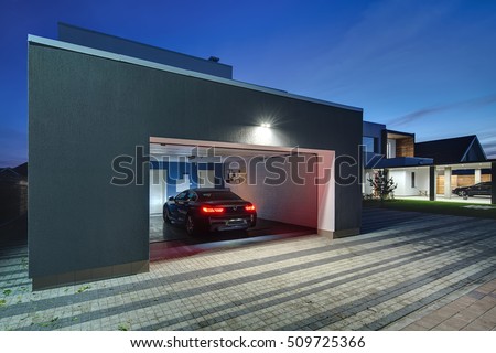 Entrance to a garage with the shining lamps in the modern country house. In the garage there is a black car with glowing parking lights. Around the house there is a tiled area. Horizontal.