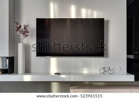 Big TV on the white wall in the room in a modern style. Under it there is a white rack with a flower in a vase, TV remote, decorations in a form of the bicycles. Closeup. Horizontal.