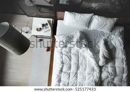 View from above at the bed with white pillows and blankets. There is a white table with book and cup, lamp with lampshade, wardrobe with glass sliding doors. On the floor there is parquet with carpet.