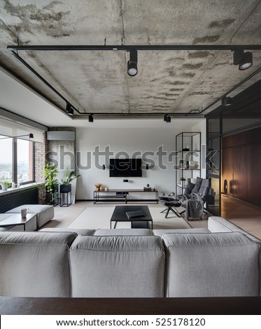 Loft style sitting-room with white, brick and concrete walls. There is sofa, tables, chair, armchair, shelves, speakers, TV and rack under it, lamps. On the floor there is a parquet and carpet.