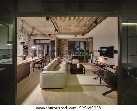 Luminous sitting-room in a loft style with different walls and a concrete ceiling. There are light sofas, wooden tables, different chairs, armchair, TV, lamps, kitchen zone with kitchen equipment.