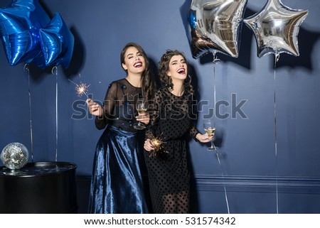 Two laughing models in trendy night dresses with glasses and sparklers in the hands on the blue wall background in the studio. There are blue and silver star-balloons, black barrel with disco ball.