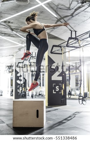 Active woman jumps above the wooden box in the gym on the background of the hanging TRX straps. She wears dark top and pants, red sneakers. Vertical.