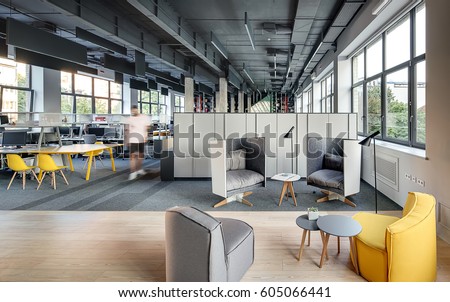 Office in a loft style with large windows, gray walls and concrete columns. There are many workplaces with computers and shelves and lockers, relax zone with armchairs with round tables and lamps.