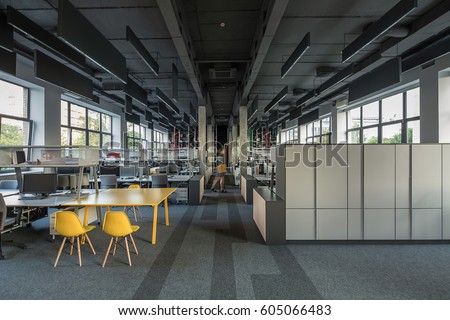 Coworking in a loft style with large windows, gray walls, concrete columns and a carpet on the floor. There are many workplaces with computers and metal reticulated shelves and lockers. Horizontal.