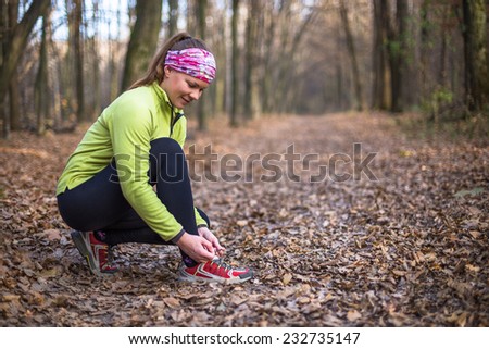 Runner woman tying running shoes outside in fall. Beautiful young fitness model smiling happy in casual jogging clothing.
