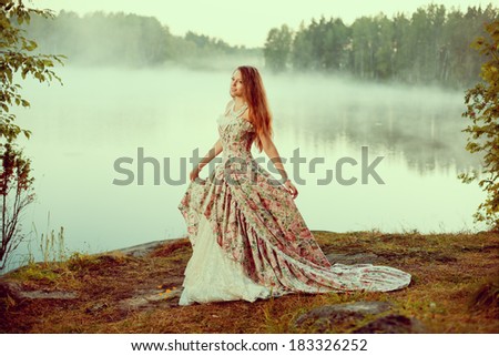 Luxury woman in a forest in a long vintage dress near the lake. Girl meets sunrise in summer forest