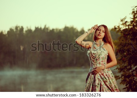 Luxury woman in a forest in a long vintage dress near the lake. Girl meets sunrise in summer forest