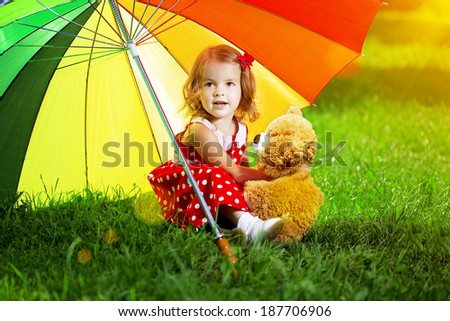 Happy smiling little girl with a rainbow umbrella in park. Child playing on green meadow. Smiling kid. Portrait of baby outdoor and teddy bear