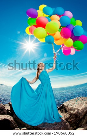 Luxury fashion stylish woman with balloons in hand on the beach against the sky and the sun in long dress