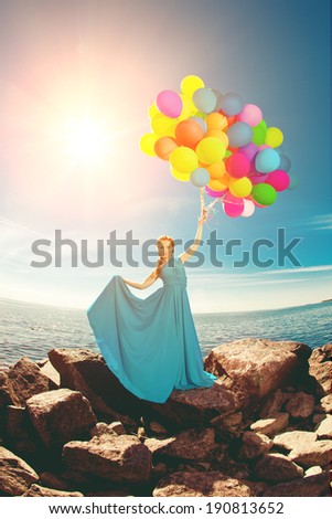 Luxury fashion stylish woman with balloons in hand on the beach against the sky and the sun in long dress