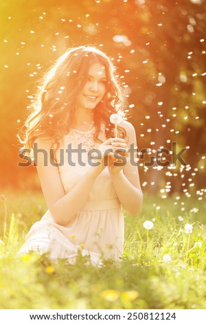 Young spring fashion woman blowing dandelion in spring garden. Springtime. Trendy girl at sunset in spring landscape background. Allergic to pollen of flowers. Spring allergy