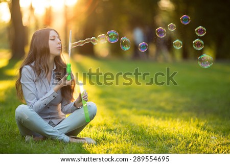Young fashion casual woman blow bubbles in sunset in park Springtime Summertime Trendy girl sitting on  grass in  sunset Spring landscape background.  Sun rays Summer scene Sunlight background Backlit