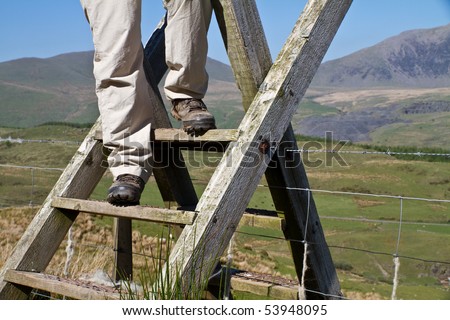 Hill walker climbing a ladder over a fence on a sunny day in Snowdonia with Snowden in the background