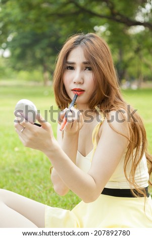 Woman applying lipstick. Asian woman with long hair sitting on the grass in the park. In applying lipstick