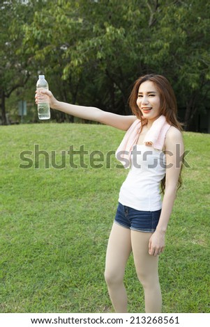 Female breaks standing and holding a bottle of water. On the grass in the public park. While jogging