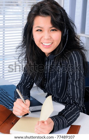 Pretty Chinese Female Call Center Agent Wearing A Telephone Headset With Long Hair Sat At Her Office Desk