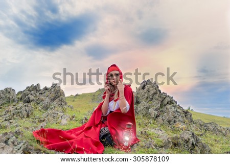 Beautiful blonde woman in old-fashioned dress and red cloak sitting on the rocks