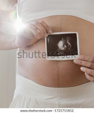 Pregnant mid adult woman holding ultrasound scan over belly, close up