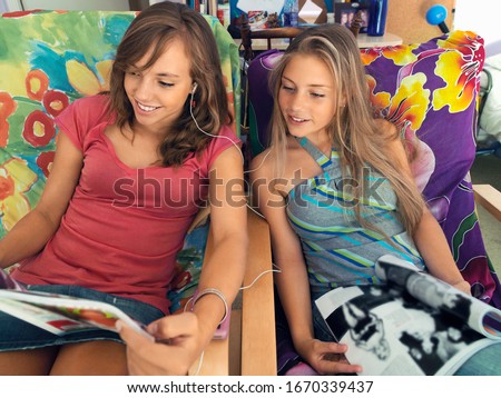 Teenage girl friends reading magazines in chairs at home listening to music on MP3 player sharing headphones