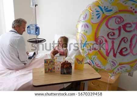 Girl recovering in hospital bed talking to doctor with get well balloon and cards in foreground