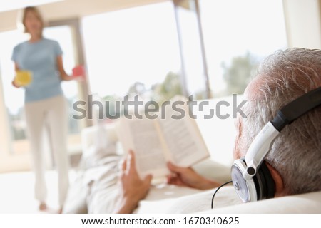 Senior man lying on sofa listening to music on headphones reading book
