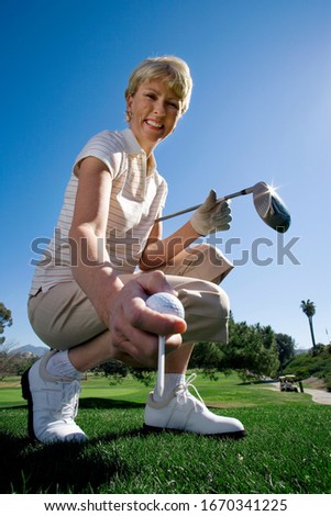 Smiling mature woman with club placing golf ball on tee at camera