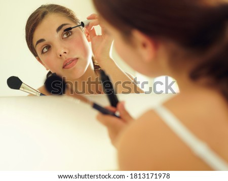 Over the shoulder view of a teenage girl looking at reflection in bathroom mirror and applying mascara.