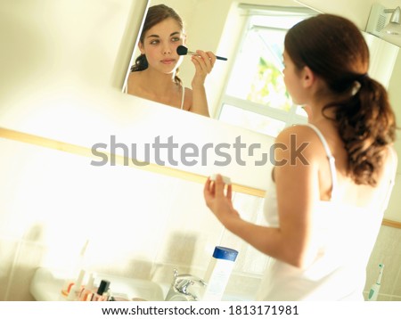 Angled shot of a teenage girl looking at reflection in bathroom mirror and applying makeup with a brush.