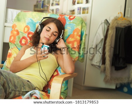 A female teenager resting on a chair while listening to music through wired headphones.