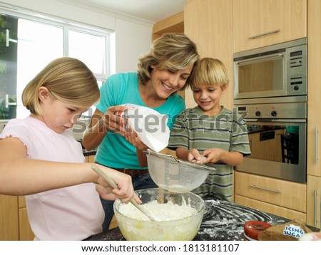 A mother helping her kids in the kitchen as they prepare something in a glass bowl using a metallic mesh.