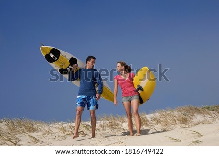 Full length of a young happy couple carrying yellow surfboards on beach over sand dunes.