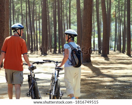 Three quarter rear view of a senior couple in cycling helmets walking side by side through wood with their bicycles with copy space.