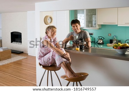 Wide shot of a happy couple in pajamas sitting at kitchen table with man drinking mug of coffee and woman with feet up on stool.