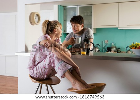 A couple in pajamas sitting at kitchen table with man drinking mug of coffee and woman with feet up on stool talk away.