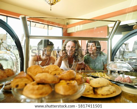 Portrait of three young women staring at pastries on display in cafe with one pointing as them seen through glass.