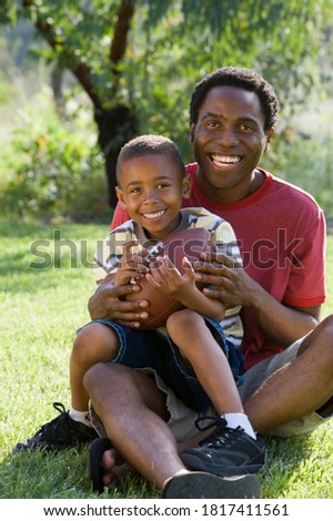 Father and son sitting on a grassy ground with a rugby ball in the hand.