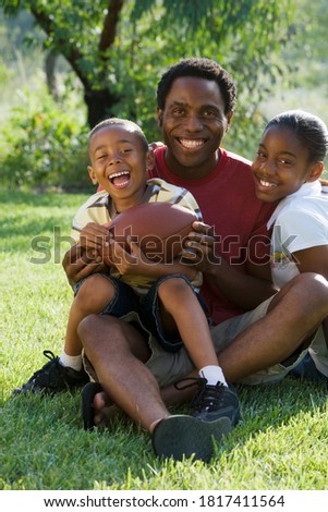 Father, son and daughter sitting on a grassy ground with the son holding a rugby ball in his hand.