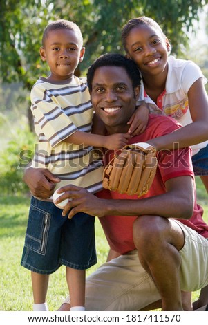 Father, son and daughter sitting on a grassy ground with the daughter wearing a baseball glove and the father holding a baseball.
