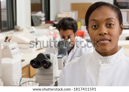 Female doctor looking at the camera with a male doctor looking through a microscope in the background.