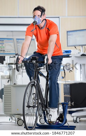 Senior man working out on an exercise bike in a laboratory while a scientist is recording his movements on a digital tablet
