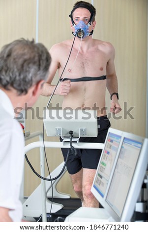 Young man running on a treadmill while a sports scientist is recording his movements on a computer using the wired mask