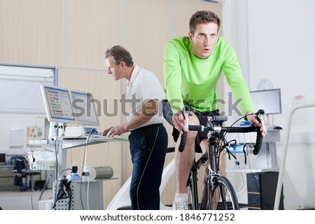 Wide shot of a sports scientist monitoring exercising data of a cyclist working out on an exercise bike