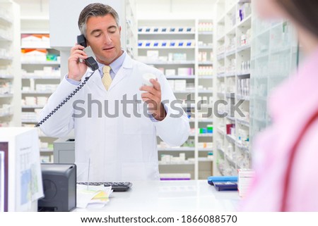 Pharmacist reading the label on a medicine pot and speaking on the phone at a pharmacy counter with a customer in the foreground.