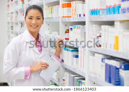 Portrait shot of a pharmacist with a prescription standing next to a pharmacy shelf and smiling at the camera.