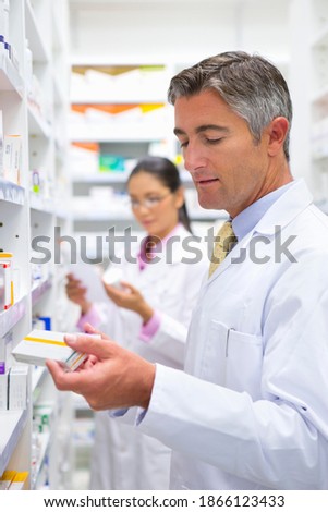 Vertical shot of a male pharmacist reading the label on a medicine boxes at a pharmacy with a colleague in the background.